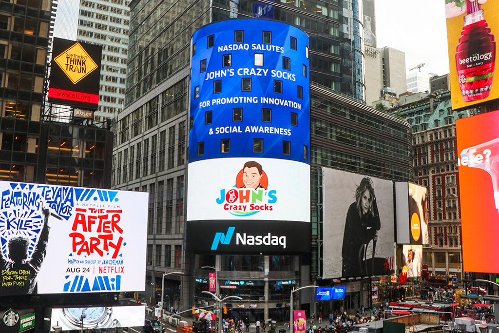 A Nasdaq billboard in Times Square honors John's Crazy Socks for promoting innovation and social awareness, featuring the brand's logo and message.