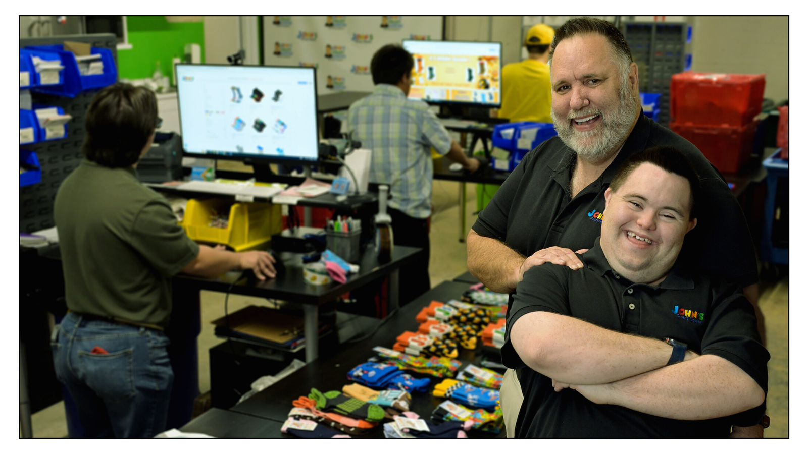 John and Mark Cronin of John's Crazy Socks smile in their vibrant, inclusive workspace as team members package socks in the background.
